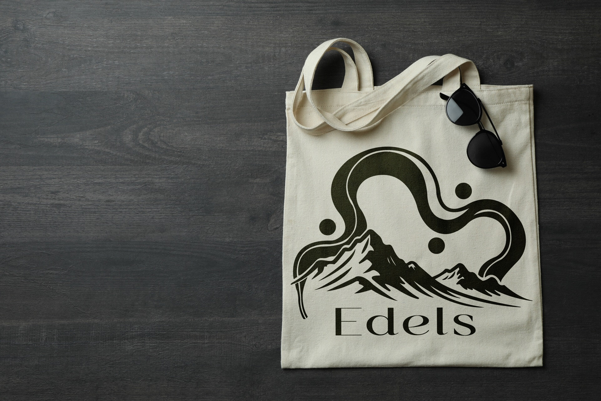 Handlenett med "Edels"-logo og fjellmotiv liggende på mørkt trebord ved solbriller