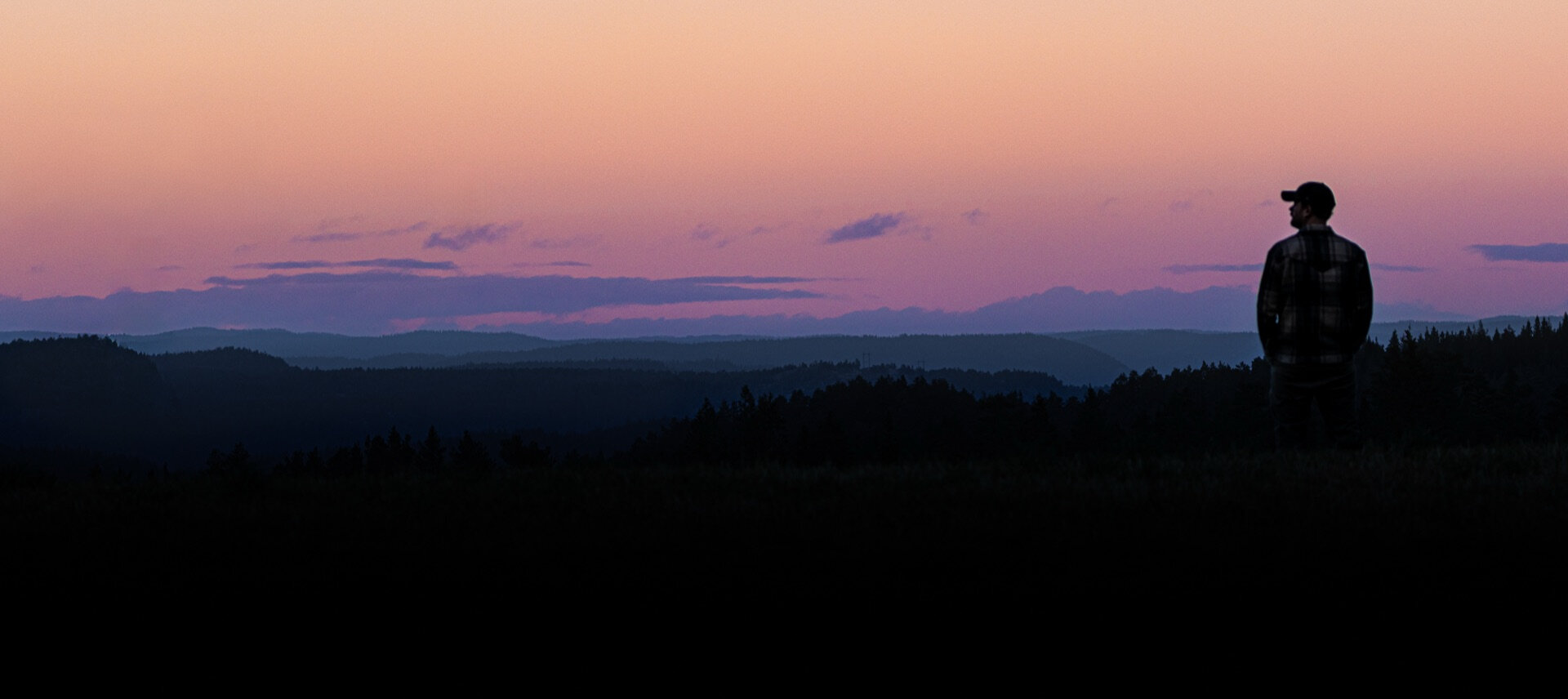 Silhuett av person som ser utover skogkledd dal ved solnedgang